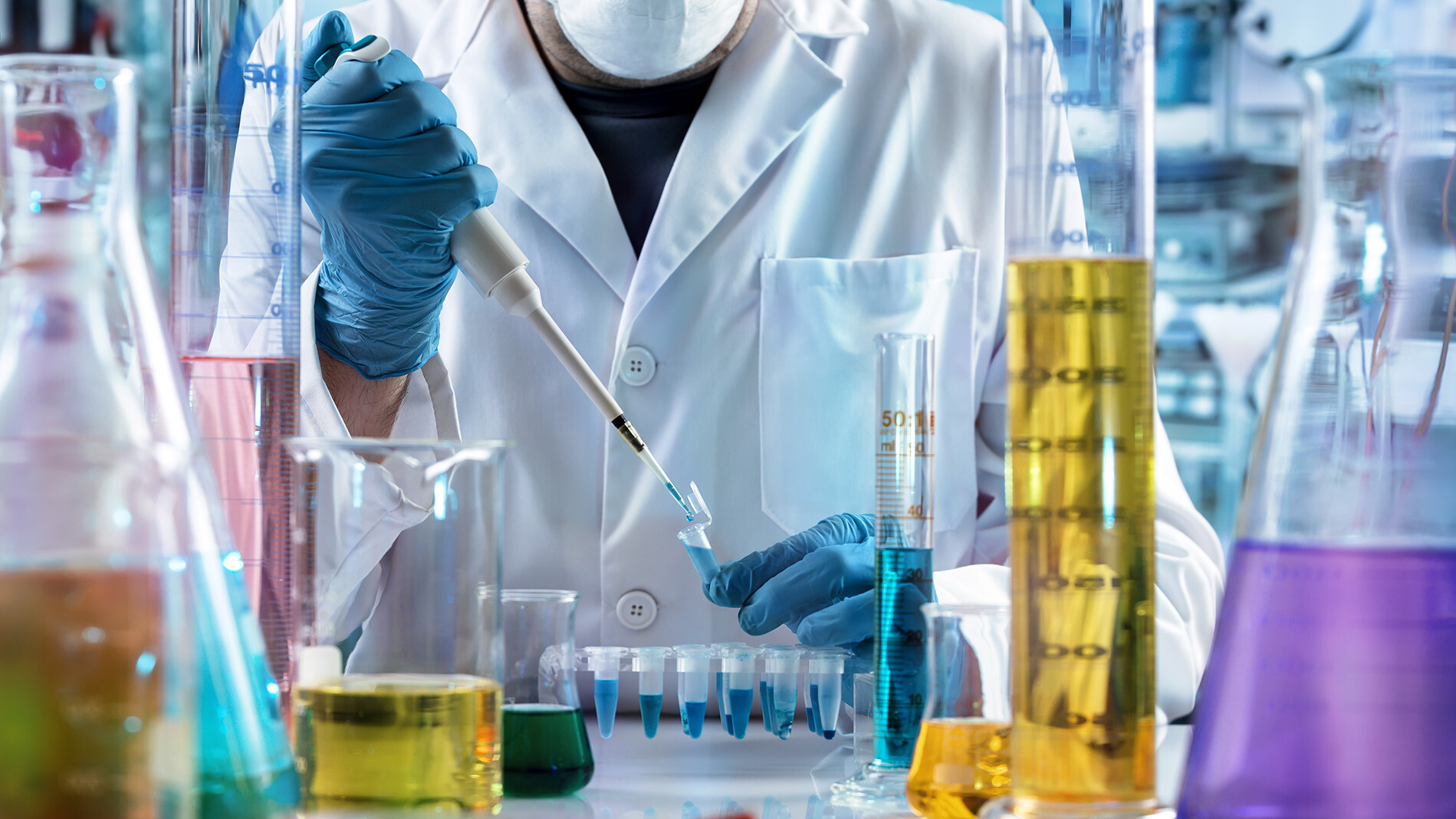 a tech wearing a protective mask and white lab coat using pipettes 